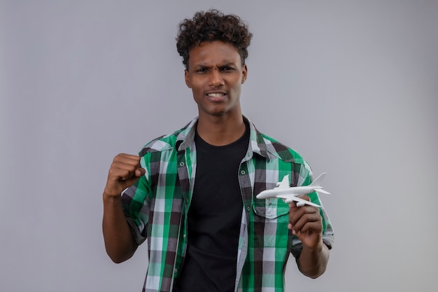 Homem jovem viajante afro-americano segurando um avião de brinquedo, levantando o punho, saiu e feliz levantando o punho, regozijando-se com seu sucesso em pé sobre um fundo branco