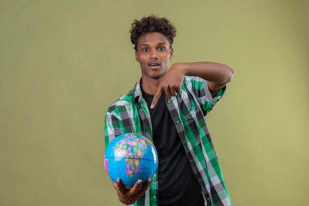 Homem jovem viajante afro-americano segurando um globo apontando com o dedo e olhando para a câmera saiu