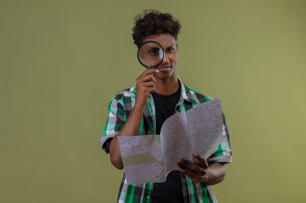 Homem jovem viajante afro-americano segurando um mapa, olhando para a câmera através de uma lupa, sorrindo em pé sobre um fundo verde