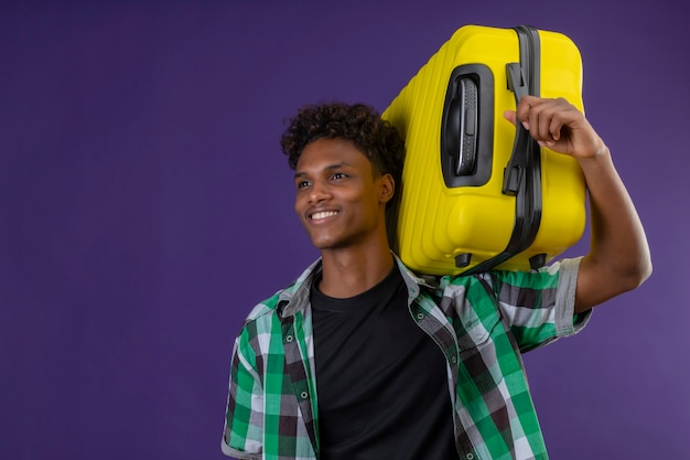 Homem jovem viajante afro-americano segurando uma mala, olhando para o lado, sorrindo, feliz e positivo em pé sobre o fundo roxo