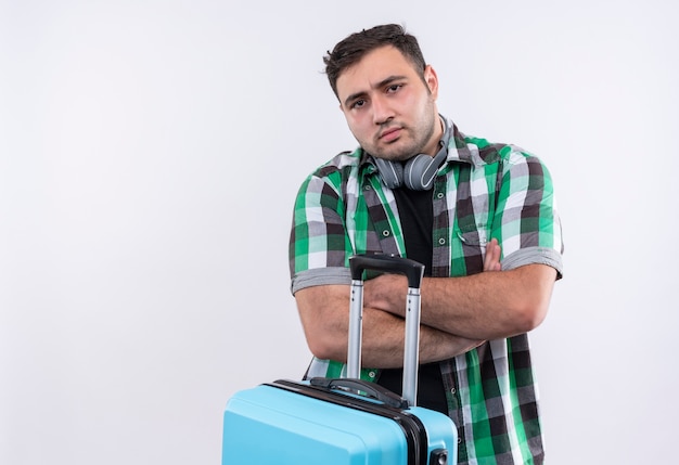 Foto grátis homem jovem viajante com camisa xadrez segurando uma mala e rosto carrancudo em pé sobre uma parede branca