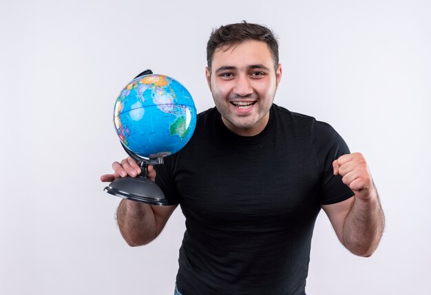 Homem jovem viajante com camiseta preta segurando um globo cerrando o punho feliz e positivo sorrindo amplamente em pé sobre uma parede branca