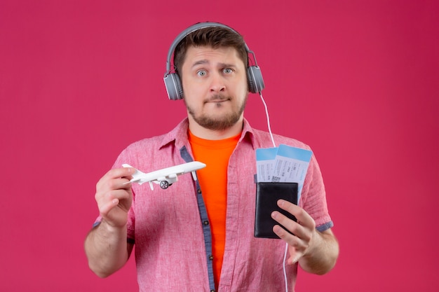 Homem jovem viajante com fones de ouvido segurando o avião de brinquedo e passagens aéreas, alegre e feliz olhando para a câmera, confuso e preocupado em pé sobre um fundo rosa