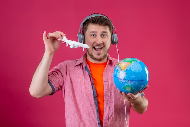 Homem jovem viajante com fones de ouvido segurando um globo e um avião de brinquedo alegre e feliz olhando para a câmera em pé sobre o fundo rosa