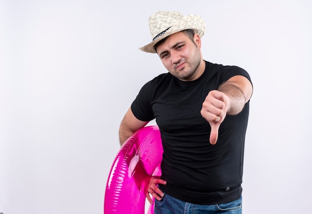 Foto grátis homem jovem viajante descontente com uma camiseta preta e um chapéu de verão segurando um anel inflável e mostrando os polegares para baixo em pé sobre uma parede branca