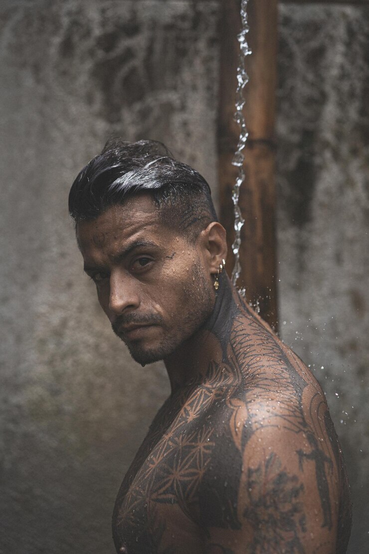 Foto grátis homem latino-americano musculoso sem camisa com tatuagens em pé tomando banho outloodrs após treino no fundo de folhas tropicais. chuveiro na villa privada.