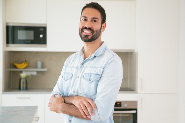 Homem latino bonito de cabelos escuros feliz posando com os braços cruzados na cozinha