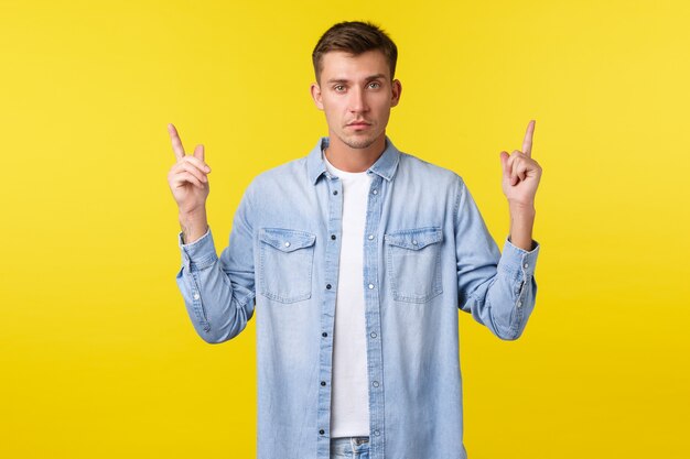 Homem loiro bonito sério confiante apontando os dedos para cima e olhando a câmera determinada, fornecer informações sobre o evento, demonstrar o anúncio de banner, fundo amarelo permanente.