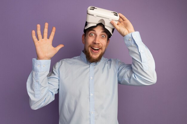 Homem louro bonito e surpreso segurando um fone de ouvido vr e se levantando com a mão erguida na parede roxa