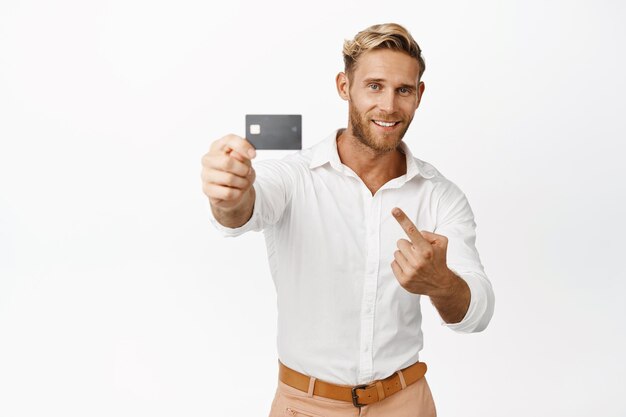 Homem macho bonito apontando e mostrando produto bancário de cartão de crédito anunciando oferta promocional de grande venda em pé sobre fundo branco