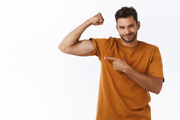 Foto grátis homem macho sorridente confiante atrevido com grandes músculos orgulhosamente apontando a mão como mostrando bíceps no braço apontando para a mão e sorrindo satisfeito treino na academia incentiva comprar associação ao clube de fitness