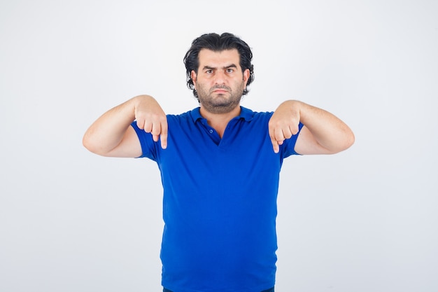 Homem maduro, apontando para baixo com os dedos indicadores em t-shirt azul, olhando com raiva, vista frontal.