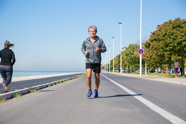 Homem maduro cansado focado em roupas esportivas, correndo ao longo da margem do rio lá fora. Treinamento de corredor sênior para maratona. Vista frontal. Conceito de atividade e idade