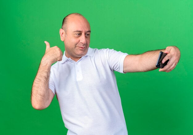 Homem maduro casual satisfeito tira uma selfie e aponta para o lado isolado no fundo verde