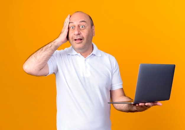 Homem maduro casual surpreso segurando laptop e colocando a mão na testa, isolado na parede amarela