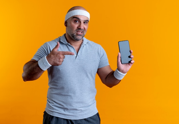 Foto grátis homem maduro e desportivo com uma bandana mostrando o smartphone apontando com o dedo para ele, parecendo confiante em pé sobre a parede laranja