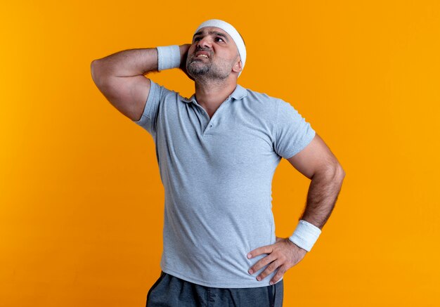 Homem maduro e desportivo com uma bandana olhando para o lado com a mão na cabeça confuso em pé sobre a parede laranja