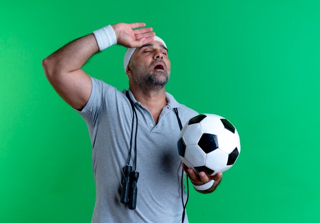 Homem maduro e desportivo com uma bandana segurando uma bola de futebol, parecendo cansado e exausto após o treino em pé sobre a parede verde