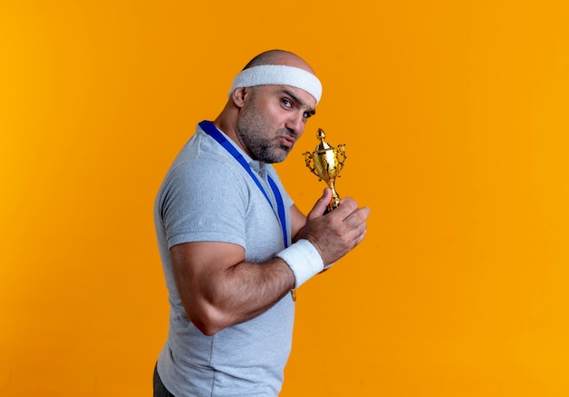 Foto grátis homem maduro e desportivo com uma faixa na cabeça e uma medalha de ouro no pescoço segurando um troféu olhando para a frente com uma cara séria em pé sobre a parede laranja