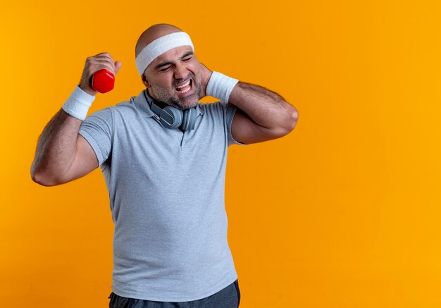 Homem maduro e desportivo com uma faixa na cabeça, malhando com halteres tocando seu pescoço, sentindo dor em pé sobre a parede laranja