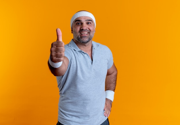 Homem maduro e desportivo com uma faixa na cabeça, olhando para a frente, sorrindo e sorrindo, mostrando os polegares em pé sobre a parede laranja