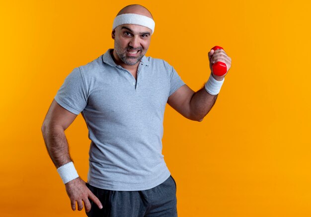 Homem maduro e desportivo com uma faixa na cabeça, segurando um haltere, parecendo confiante, fazendo exercícios em pé sobre a parede laranja