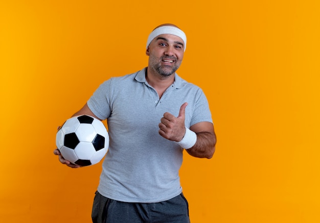 Homem maduro e desportivo com uma faixa na cabeça, segurando uma bola de futebol, olhando para a frente com um sorriso no rosto mostrando os polegares em pé sobre a parede laranja