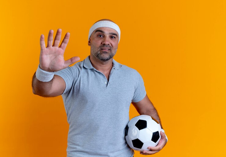 Foto grátis homem maduro e desportivo com uma faixa na cabeça, segurando uma bola de futebol, olhando para a frente, mostrando a palma da mão sobre a parede laranja