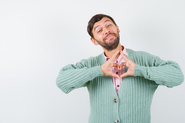 Homem maduro em camisa, casaco de lã mostrando um gesto de coração e olhando alegre, vista frontal.