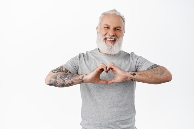 Homem maduro feliz com tatuagens rindo, mostrando o gesto de eu te amo em coração, expressar simpatia, gostar ou cuidar de alguém, em pé em uma camiseta cinza contra um fundo branco