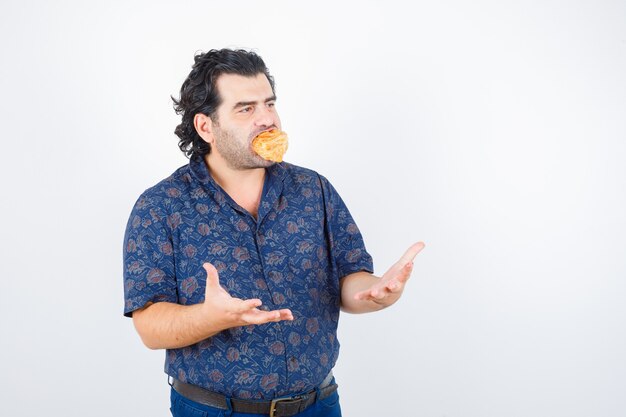 Homem maduro, segurando o produto de pastelaria na boca, mantendo as mãos de maneira agressiva na camisa e olhando sério, vista frontal.