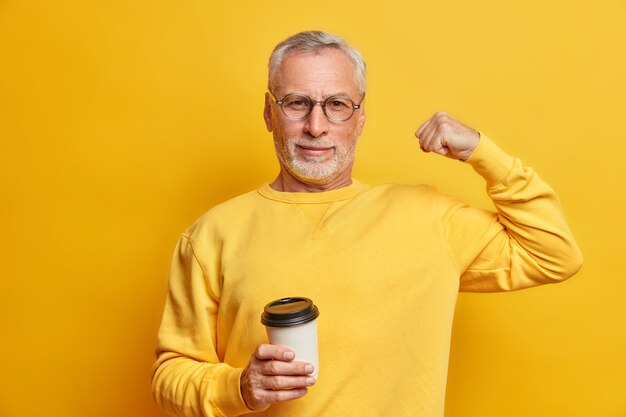 Homem maduro sério e autoconfiante levanta o braço e mostra os bíceps sendo fortes e poderosos segurando um copo de café de papel e usa um macacão casual isolado sobre a parede amarela