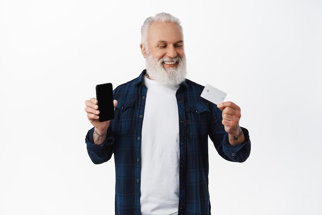 Homem maduro sorridente mostrando a tela em branco do smartphone parecendo satisfeito e feliz com o cartão de crédito do banco copyspace em pé contra o fundo branco