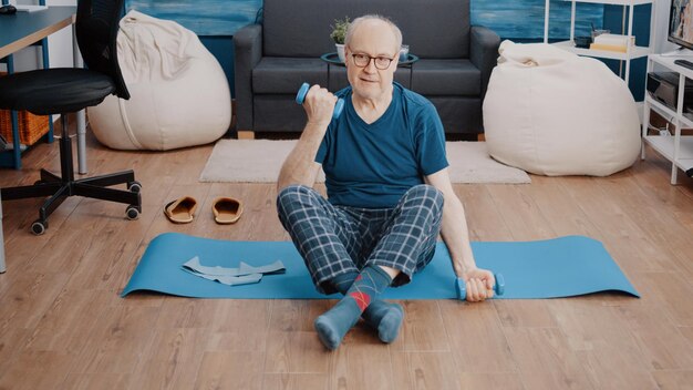 Homem mais velho sentado no tapete de ioga e levantando pesos para treinar os músculos em casa. Pensionista usando halteres para fazer exercícios físicos e esticar os braços, treinando e fazendo atividade física.