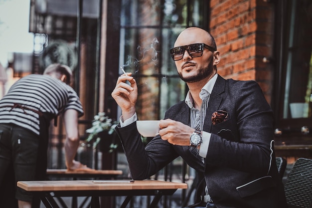 Foto grátis homem moderno pensativo está fumando um cigarro fora do coffeshop enquanto bebe sua xícara de café.