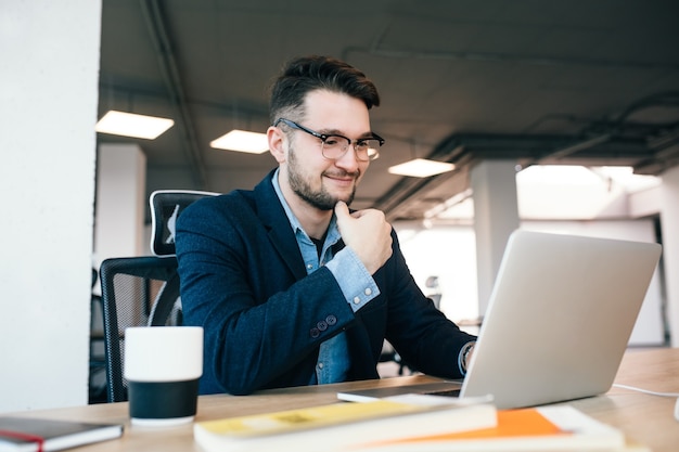 Homem moreno atraente está trabalhando com o laptop na mesa no escritório. Ele usa camisa azul com jaqueta preta. Ele está sorrindo.