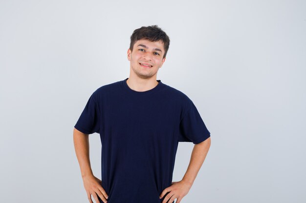 Homem moreno posando com as mãos na cintura em t-shirt e olhando alegre, vista frontal.