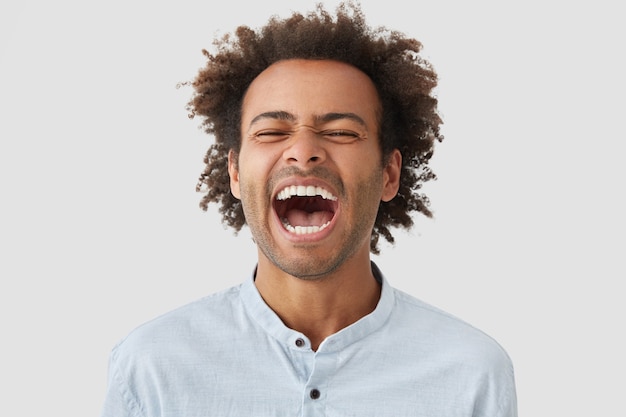 Homem muito feliz com cabelo encaracolado, ri feliz, mantém a boca bem aberta, mostra dentes brancos