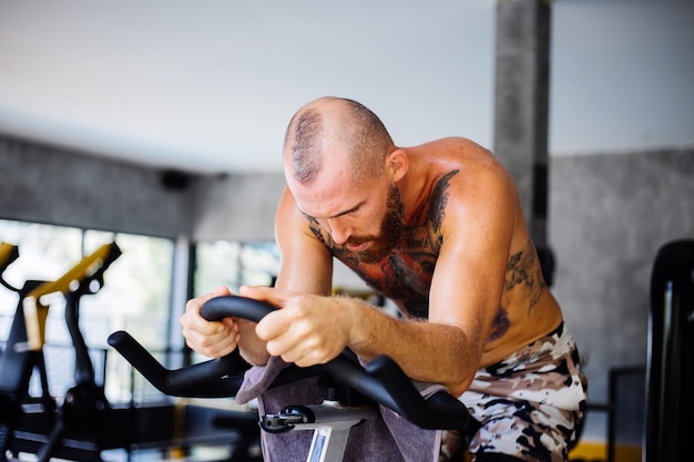 Homem musculoso e barbudo tatuado fazendo exercícios aeróbicos na bicicleta na academia perto de uma janela grande com vista para as árvores do lado de fora