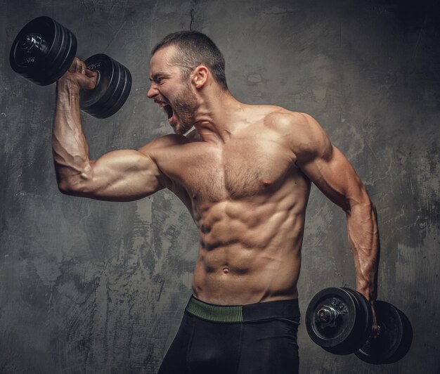 Homem musculoso gritando, sem camisa malhando com halteres em fundo cinza.