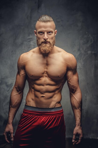 Homem musculoso sem camisa com barba posando sobre fundo cinza.