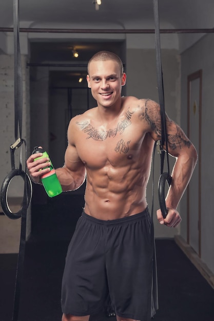 Homem musculoso sem camisa com tatuagem relaxante após treinos de anel em um clube de ginástica.