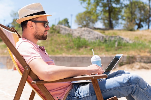 Homem na cadeira de praia, trabalhando no laptop enquanto toma uma bebida