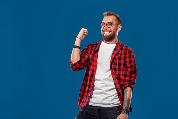 Homem na moda na camisa quadriculada, segurando os punhos em gesto de sucesso e gritando alegremente com vitória sobre fundo azul. Moderno