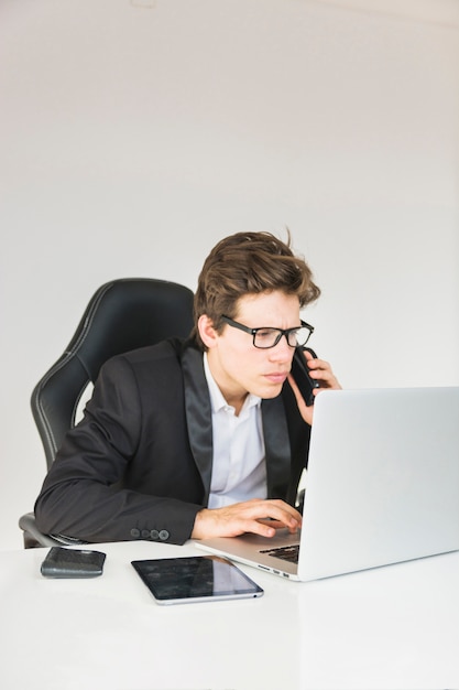 Homem negócios, falando telefone, enquanto, usando computador portátil, em, local trabalho