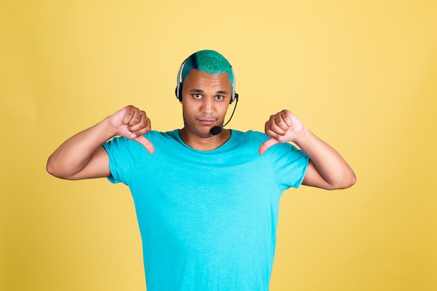 Homem negro africano casual na parede amarela cabelo azul call center trabalhador feliz operador de suporte ao cliente com fones de ouvido apontando o dedo para baixo