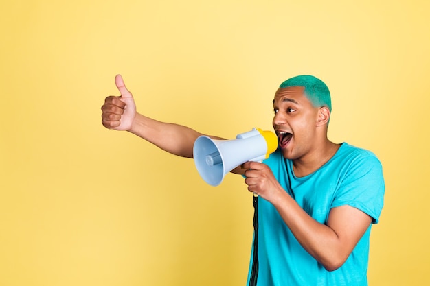 Homem negro africano casual na parede amarela de cabelo azul gritando e gritando no megafone para a esquerda mostrar o polegar para cima