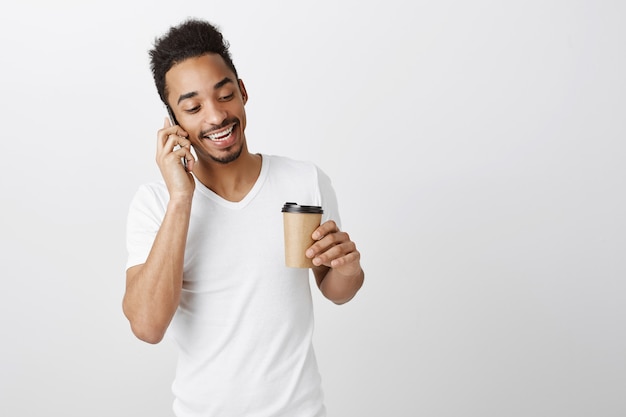 Homem negro bonito com camiseta branca falando no celular e bebendo café para viagem