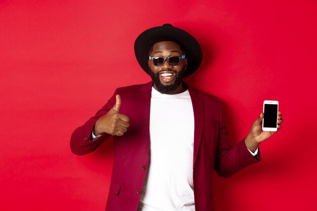 Homem negro bonito e elegante mostrando a tela do telefone e o polegar para cima para a câmera, recomendando o aplicativo da loja online, fundo vermelho