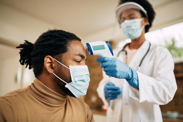 Homem negro com máscara facial medindo sua temperatura por um médico durante visita domiciliar